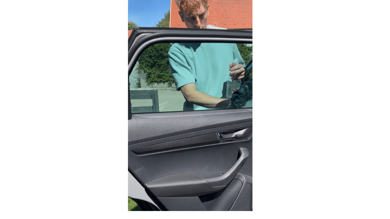Ein Mann im blauen T-Shirt reinigt ein Autofenster mit einer Sprühflasche und einem Tuch.