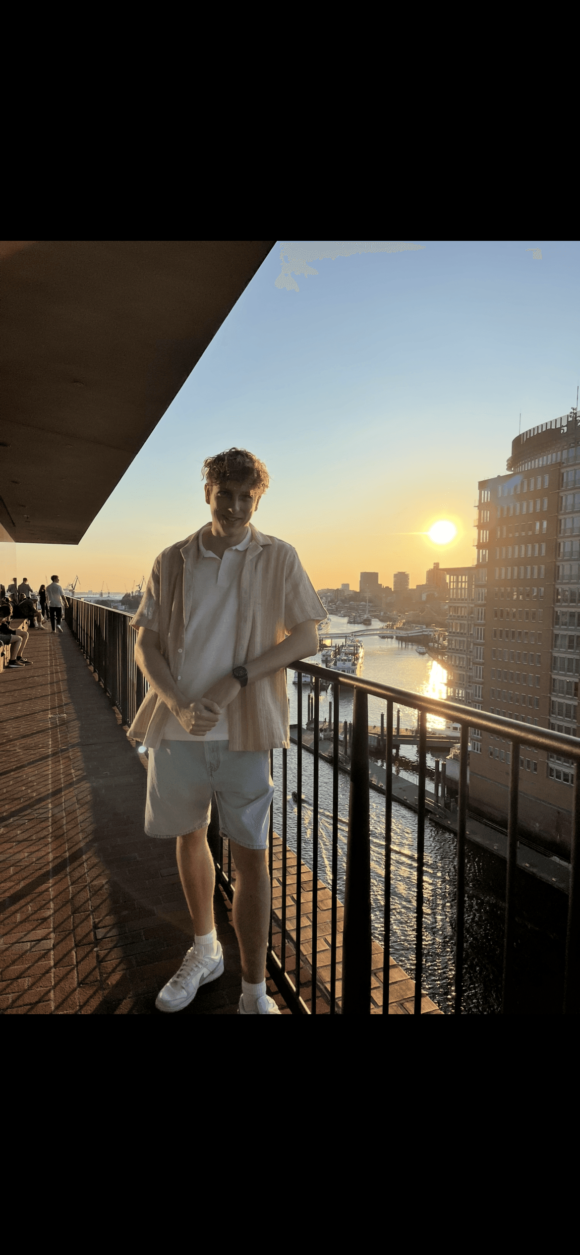 Junger Mann lächelt auf einer Aussichtsplattform vor einem Hafenbecken im warmen Licht des Sonnenuntergangs.