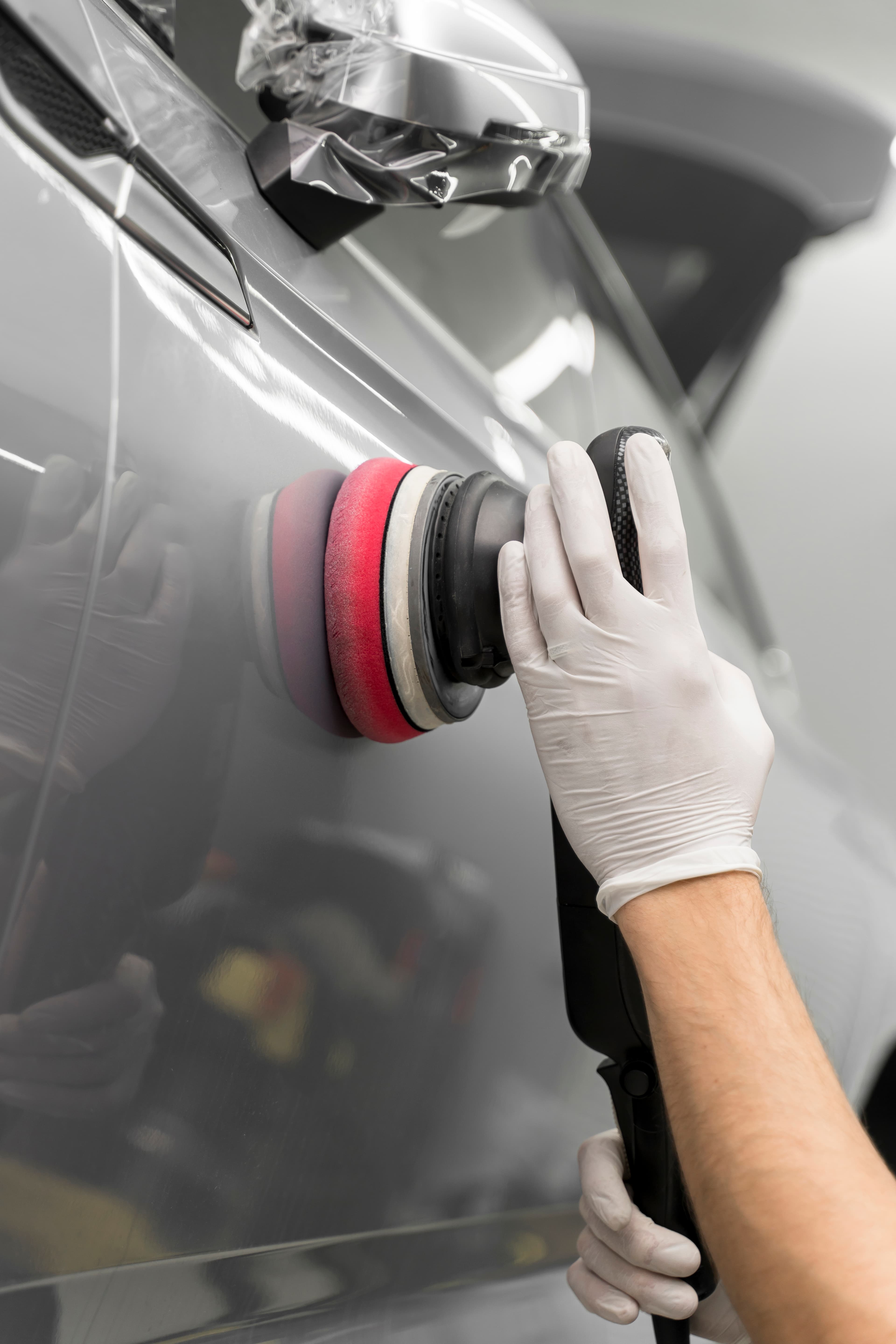 Hände in weißen Handschuhen polieren eine graue Autotür mit einer Poliermaschine und rotem Polierpad.