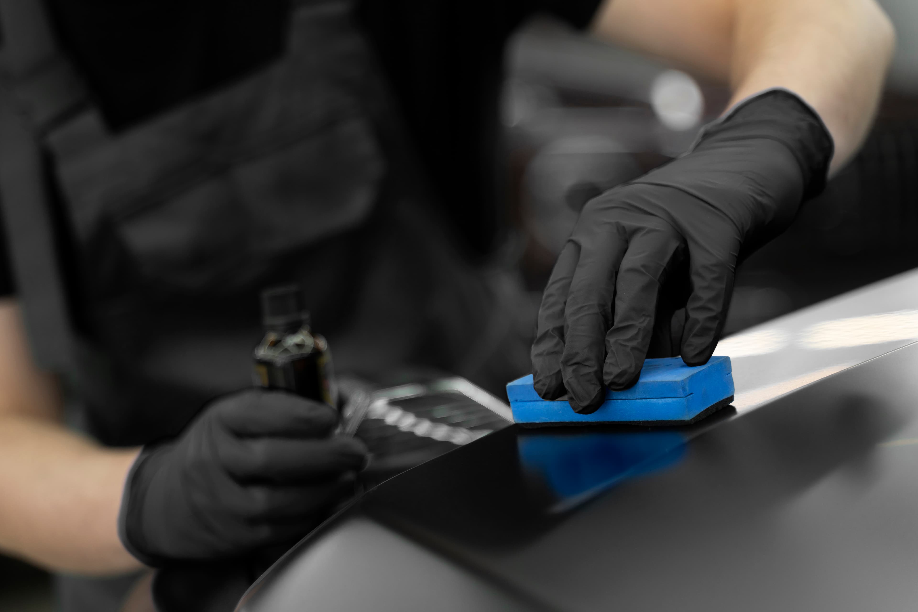 Person in schwarzen Handschuhen trägt Keramikversiegelung mit einem blauen Schwamm auf ein Auto auf.