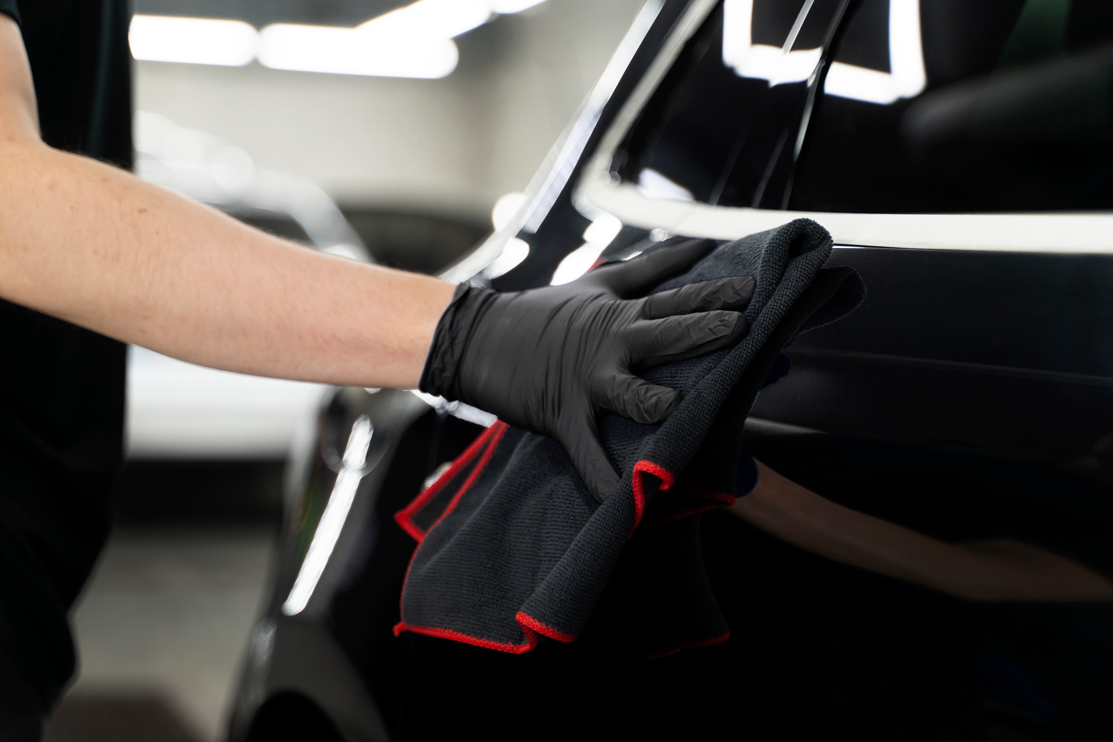 Person mit schwarzen Handschuhen poliert ein schwarzes Auto mit einem Mikrofasertuch in einer Werkstatt.