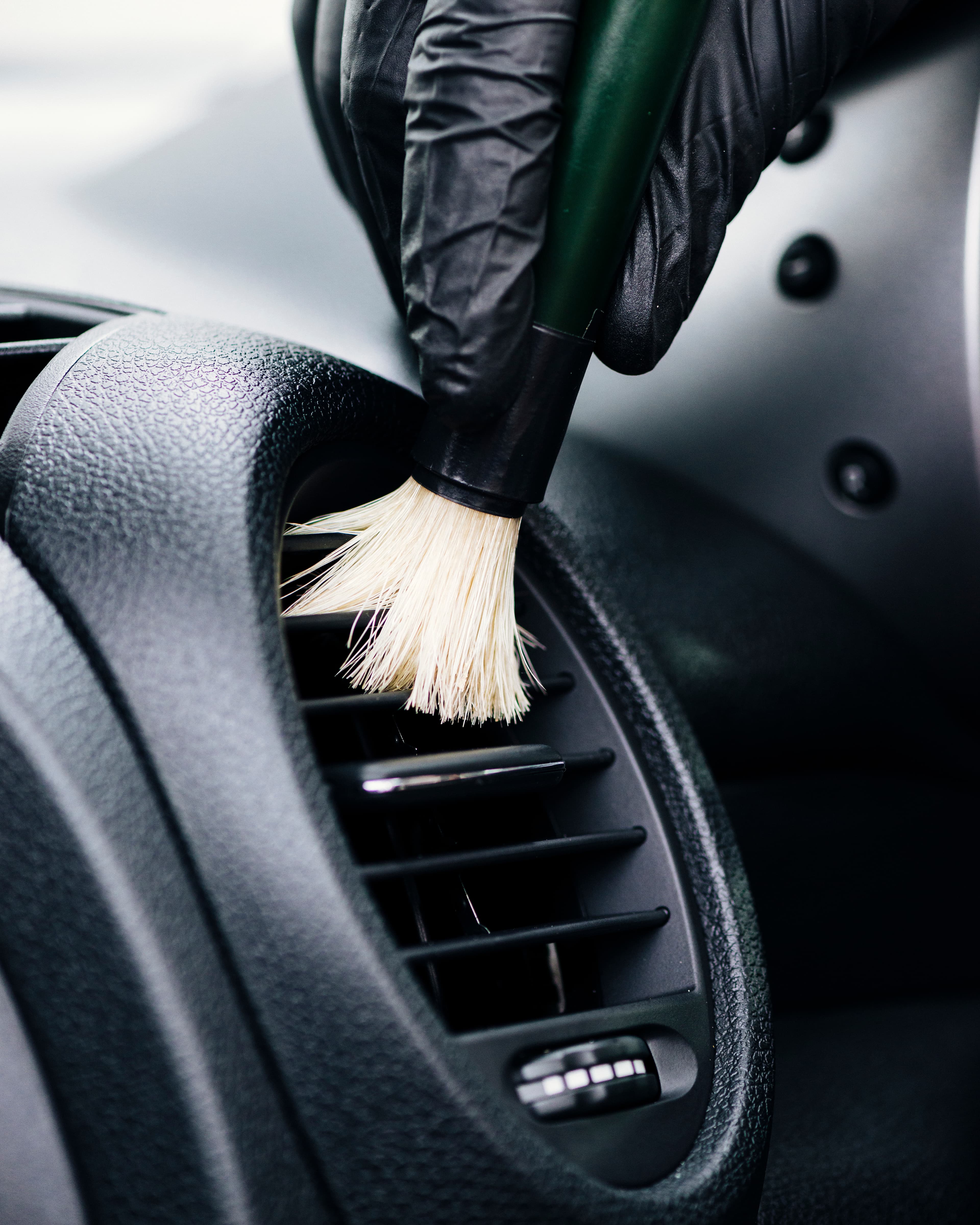 Eine behandschuhte Hand reinigt die Lüftungsschlitze eines Autos sorgfältig mit einem feinen Detailing-Pinsel.