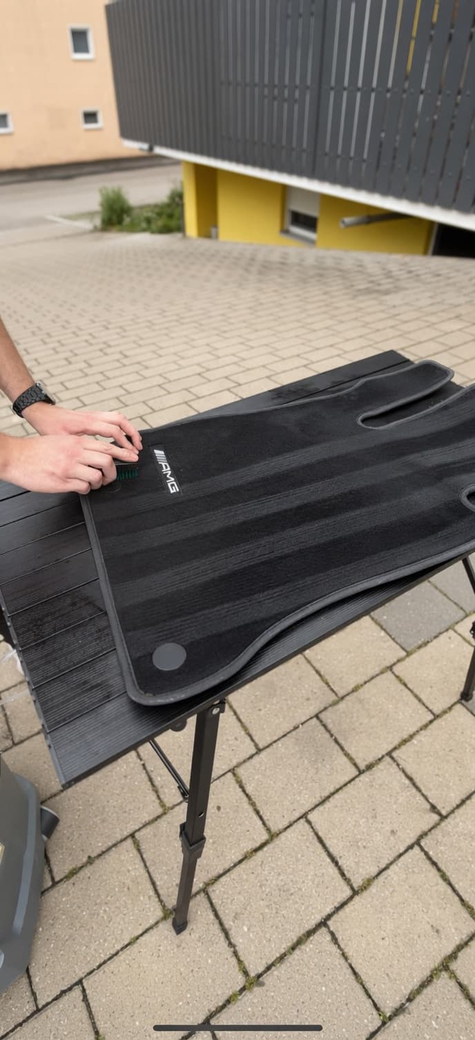 Eine Person reinigt eine schwarze AMG-Fußmatte mit einer Bürste auf einem Tisch im Freien.